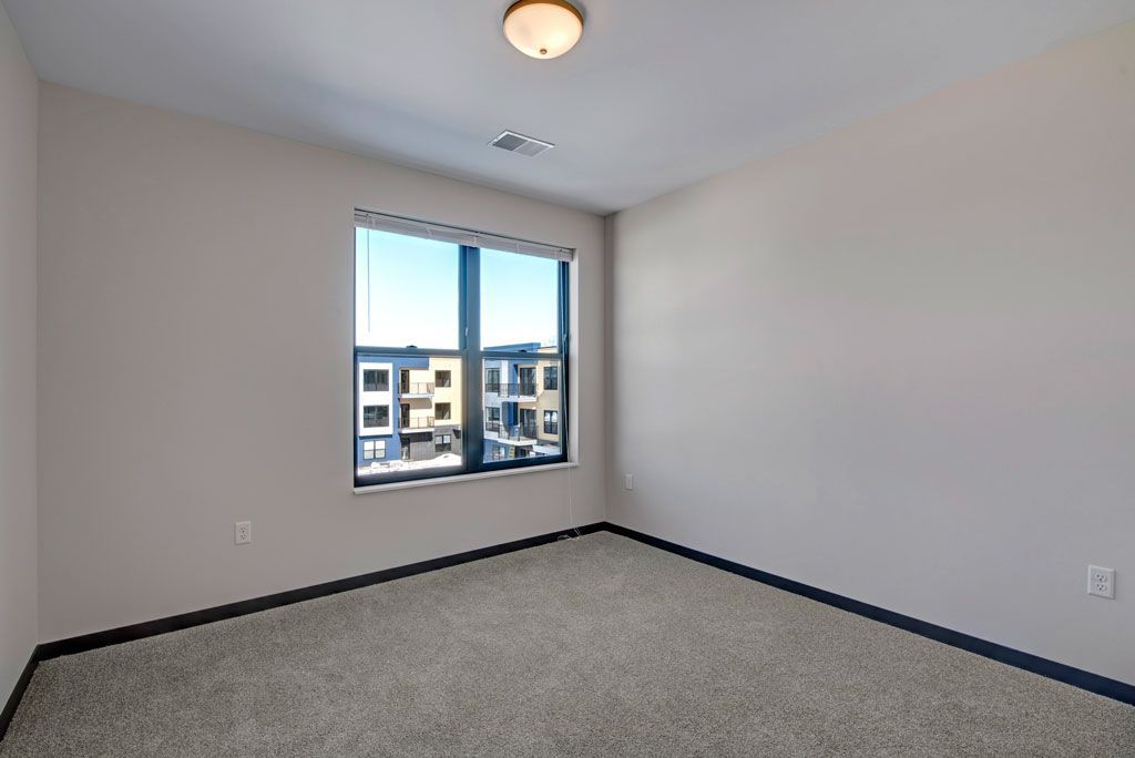 Empty bedroom with gray carpet, white walls, and a window overlooking apartment buildings.