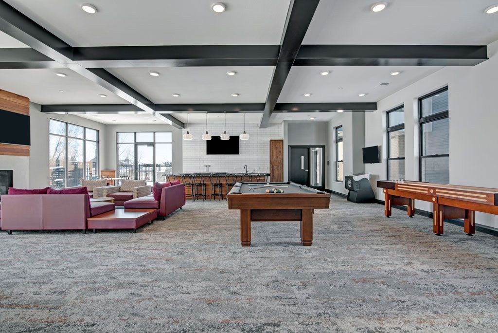 Bright modern lounge with couches, wooden tables, carpeted floor, and large windows beneath exposed beams