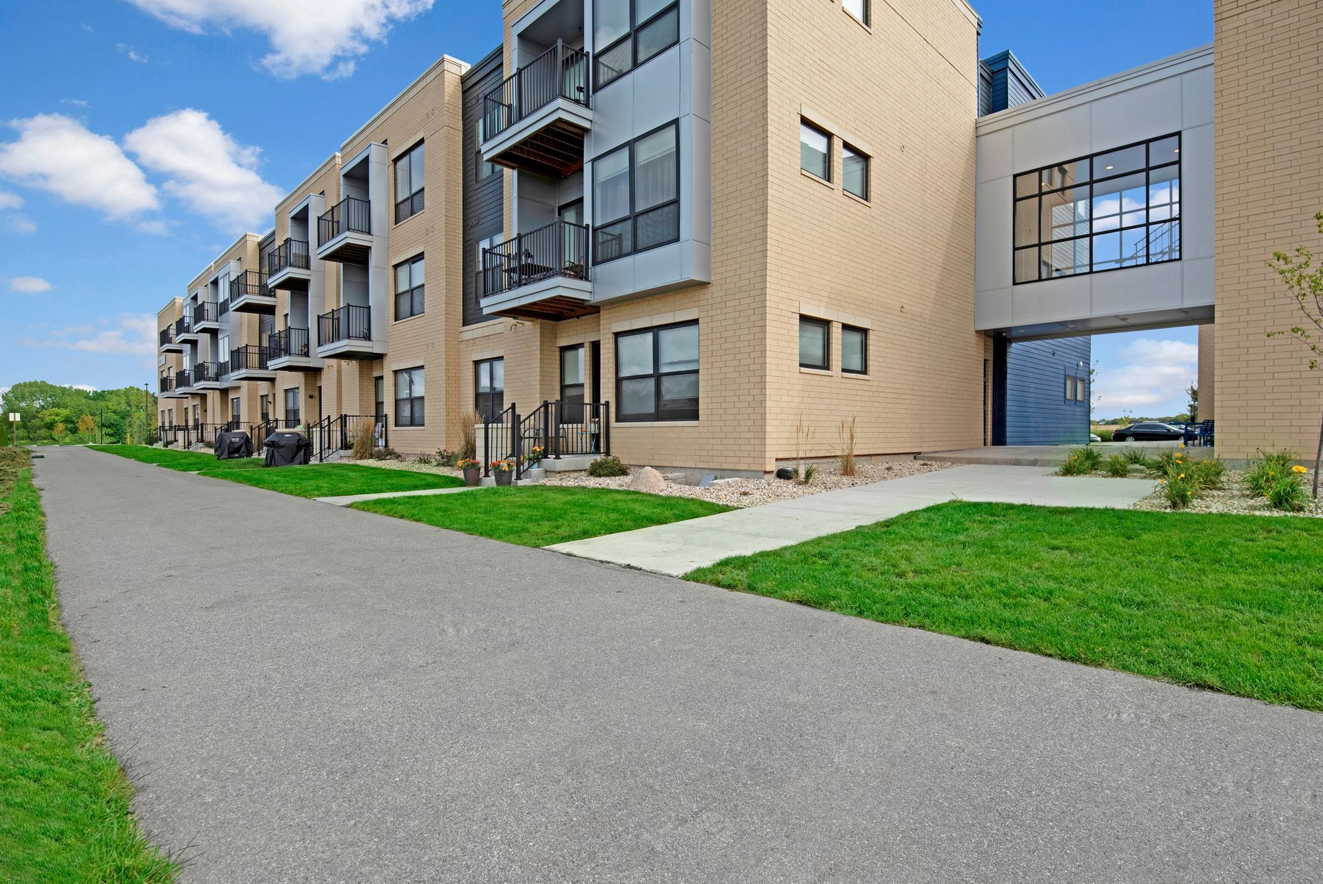 Apartment building with tan brick balconies, green lawn, and a paved walkway under a blue sky.