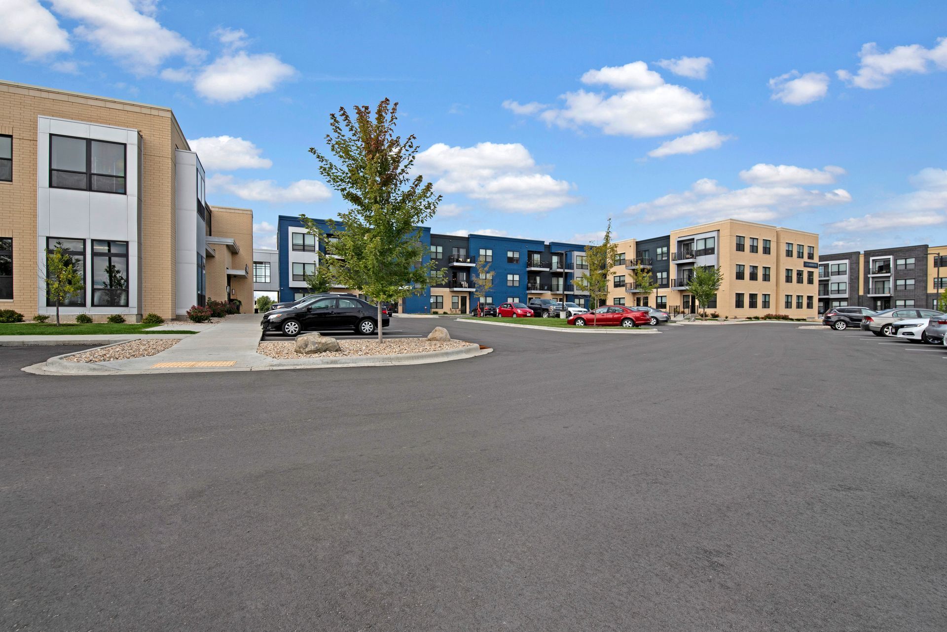 Apartment complex parking lot with modern buildings, parked cars, and a tree under a blue sky