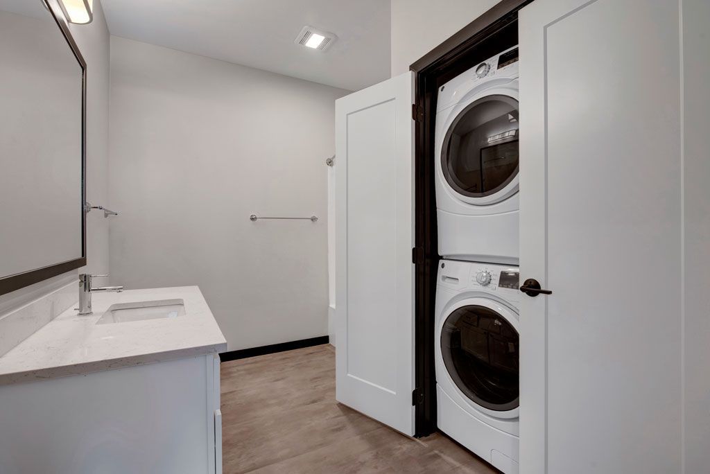 Bright laundry room with stacked washer and dryer behind white cabinet doors and a sink on the left.