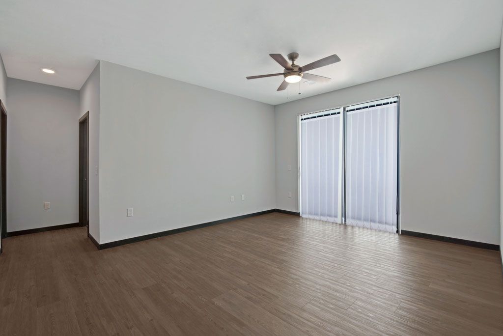 Empty bedroom with wood floors, light gray walls, ceiling fan, and sliding glass doors with blinds
