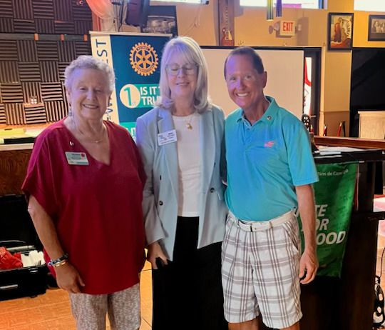Three people standing, posing for a photo at an event. A woman in red top, one in blue, and a man in a blue shirt with shorts.