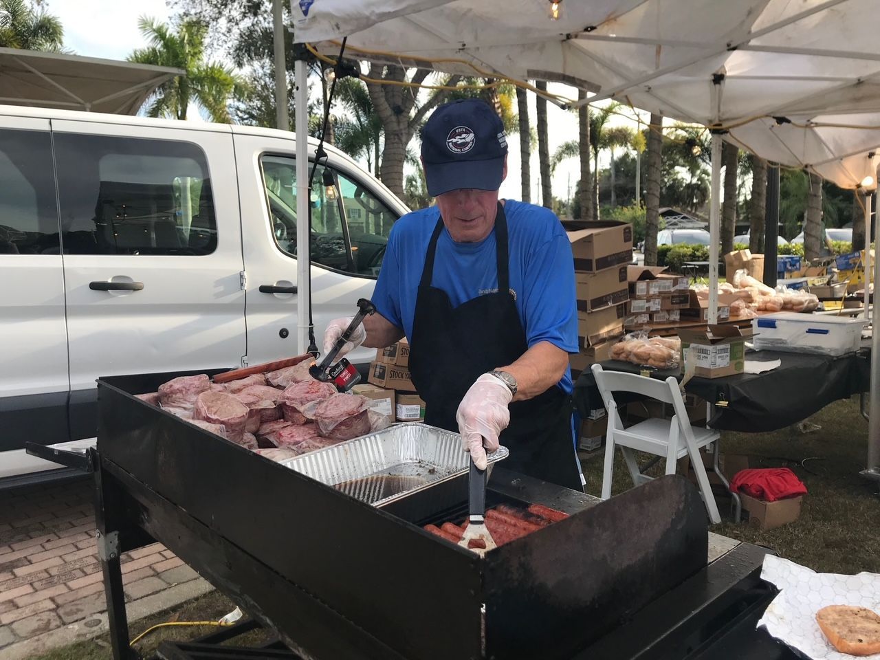 A man is cooking meat on a grill in front of a white van.