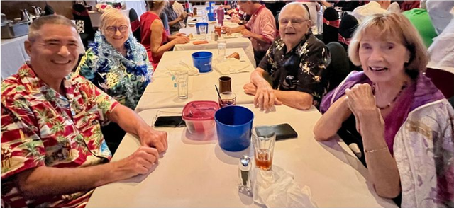 Four people smiling at a table, with food and drinks. Others seated at tables in the background.