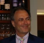 A man in a suit and tie is smiling in front of a wine shelf.