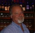 A man with a beard is standing in front of a wine shelf.
