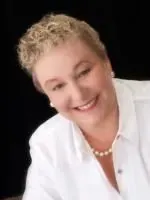 Smiling woman with short blonde hair, wearing white shirt and pearl necklace, black background.