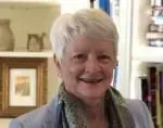 Woman with short white hair, smiling. Wearing a blue blazer and scarf, in front of a bookshelf.