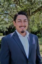 A man in a suit and white shirt is standing in front of a tree.