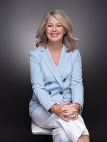 A woman in a blue jacket and white pants is sitting on a stool.