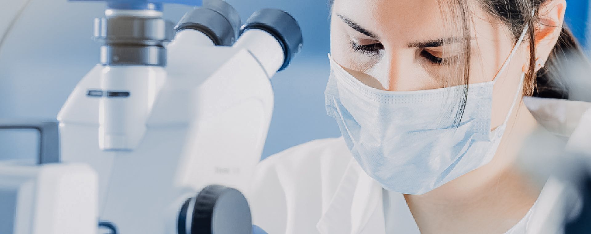 A woman wearing a mask is looking through a microscope.