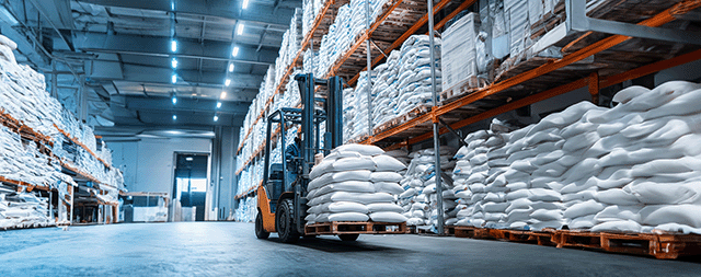 Gabelstapler in einem Lagerhaus, der Paletten mit weißen Säcken transportiert. An den Wänden stehen Regale mit Säcken.