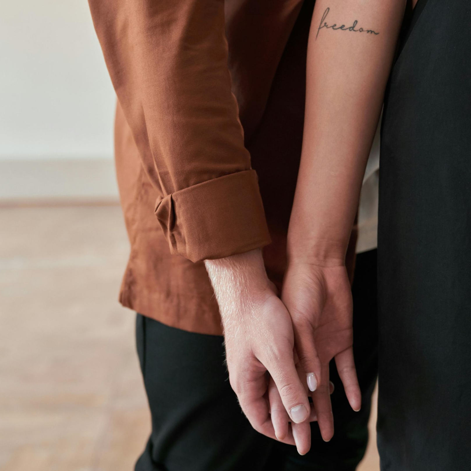 Een stel houdt elkaars hand vast; op één arm staat een tatoeage met 'vrijheid', op de andere een bruine overhemdmouw.