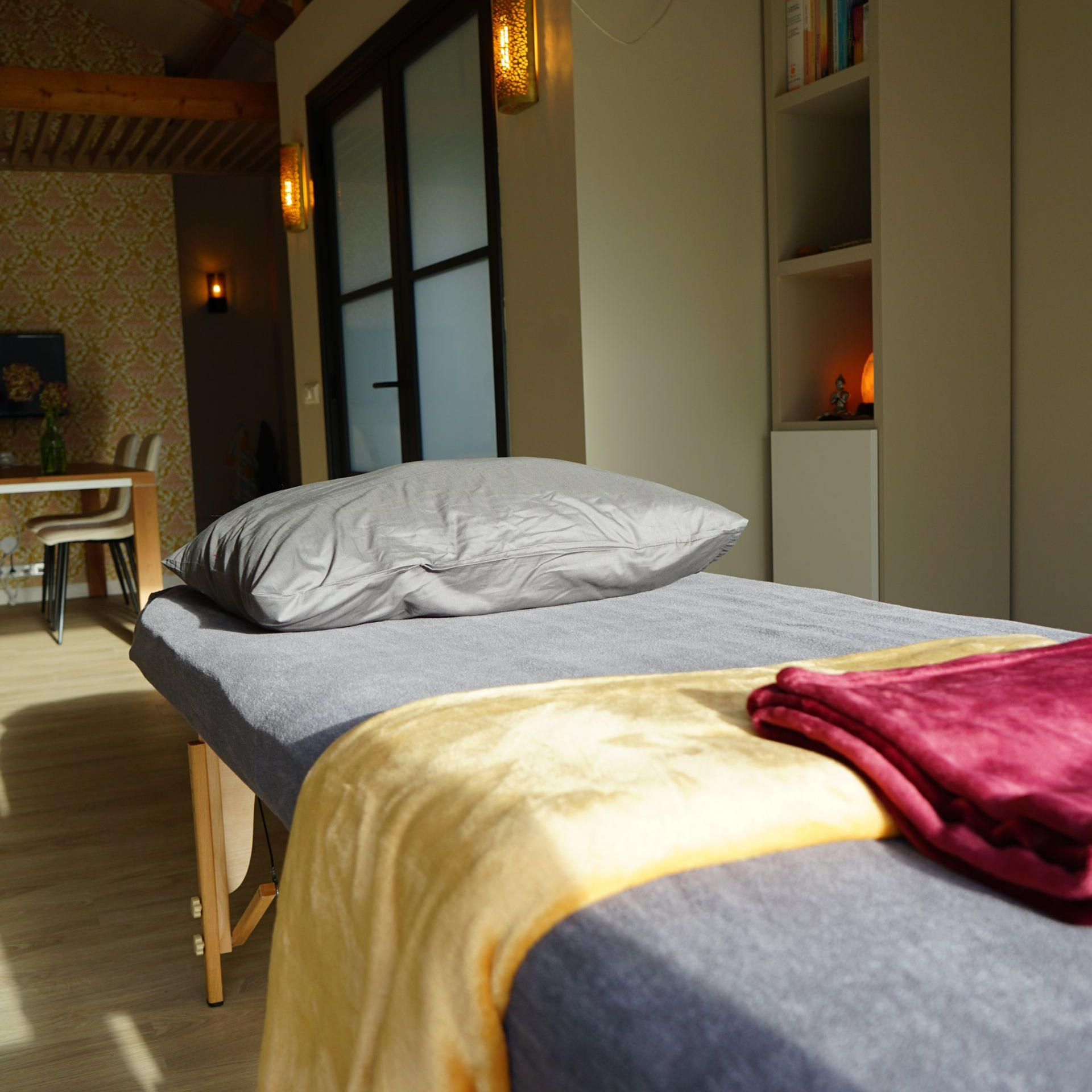 Een massagetafel in een zonnige kamer, met een grijs kussen en dekens.