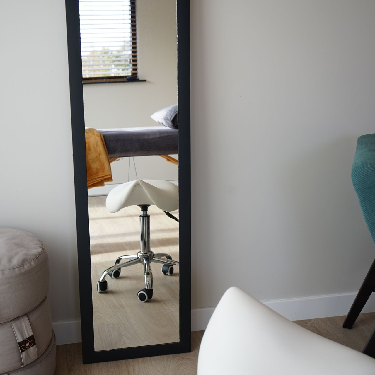 Een grote zwarte spiegel met frame in een kamer, waarin een massagetafel, een kruk en een deel van een stoel zijn afgebeeld.