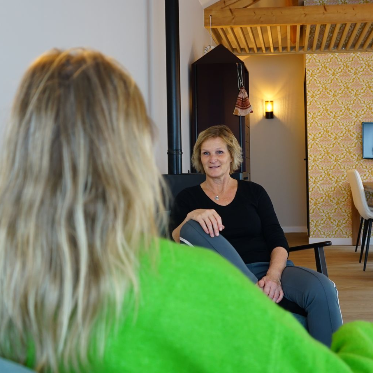 Vrouw in zwart shirt zit tegenover een andere persoon. Felgroene trui op de voorgrond, houten interieur.