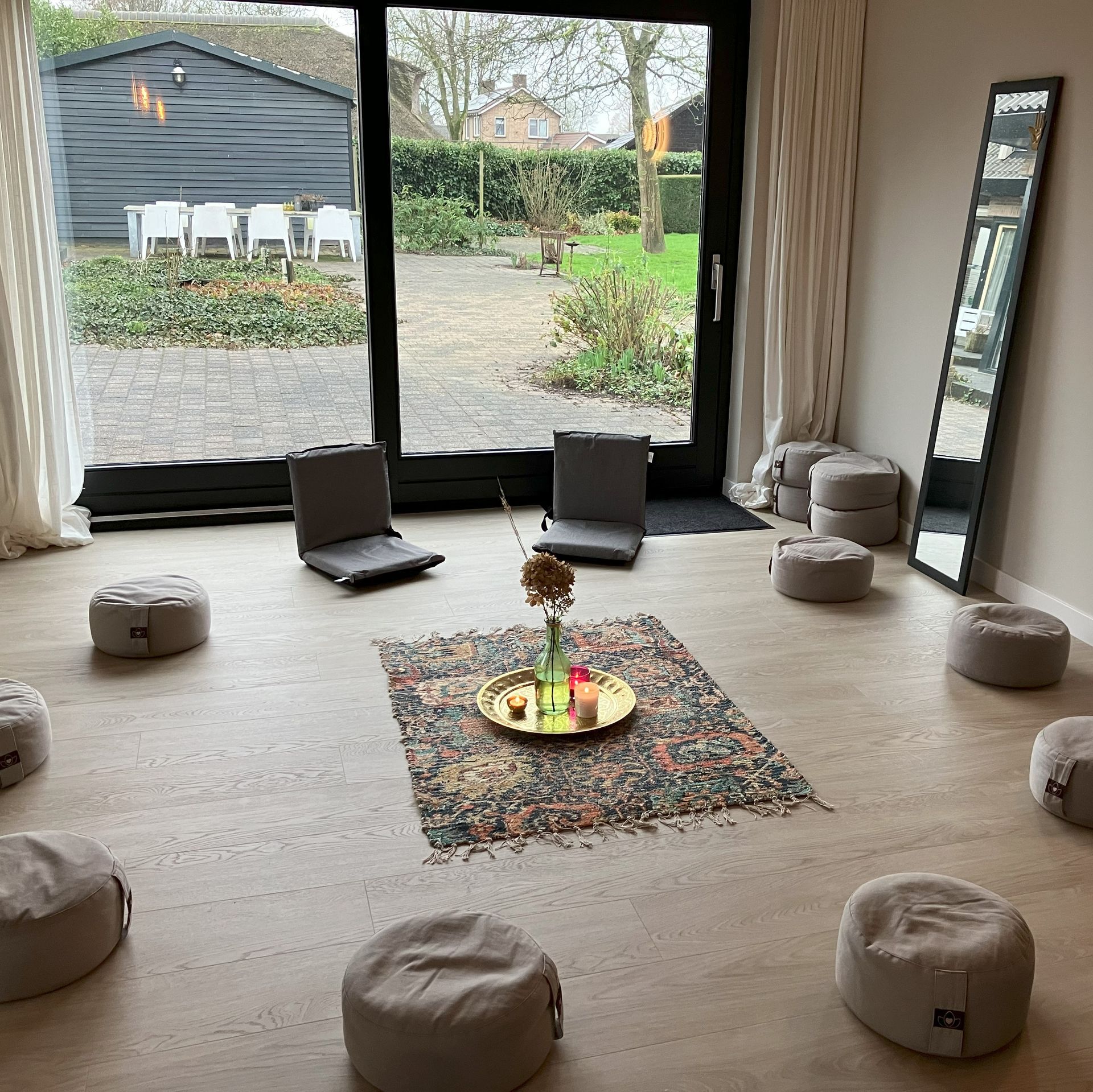 Meditatieruimte met vloerkussens, tapijt, grote spiegel en glazen deur met uitzicht op een tuin.