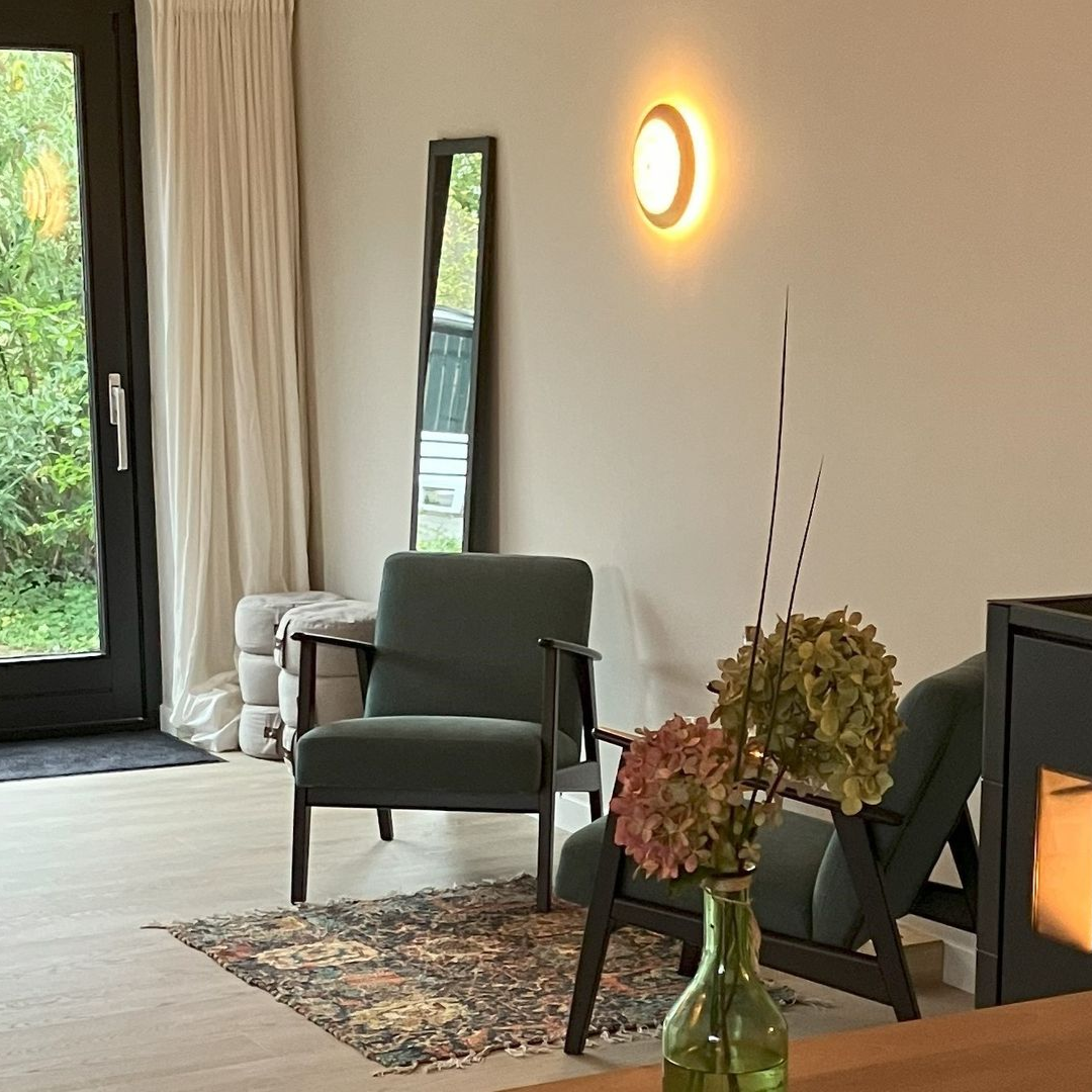 Gezellige praktijkruimte met groene fauteuils, een spiegel en decoratieve bloemen. Een wandlamp in warme tinten verlicht de kamer.