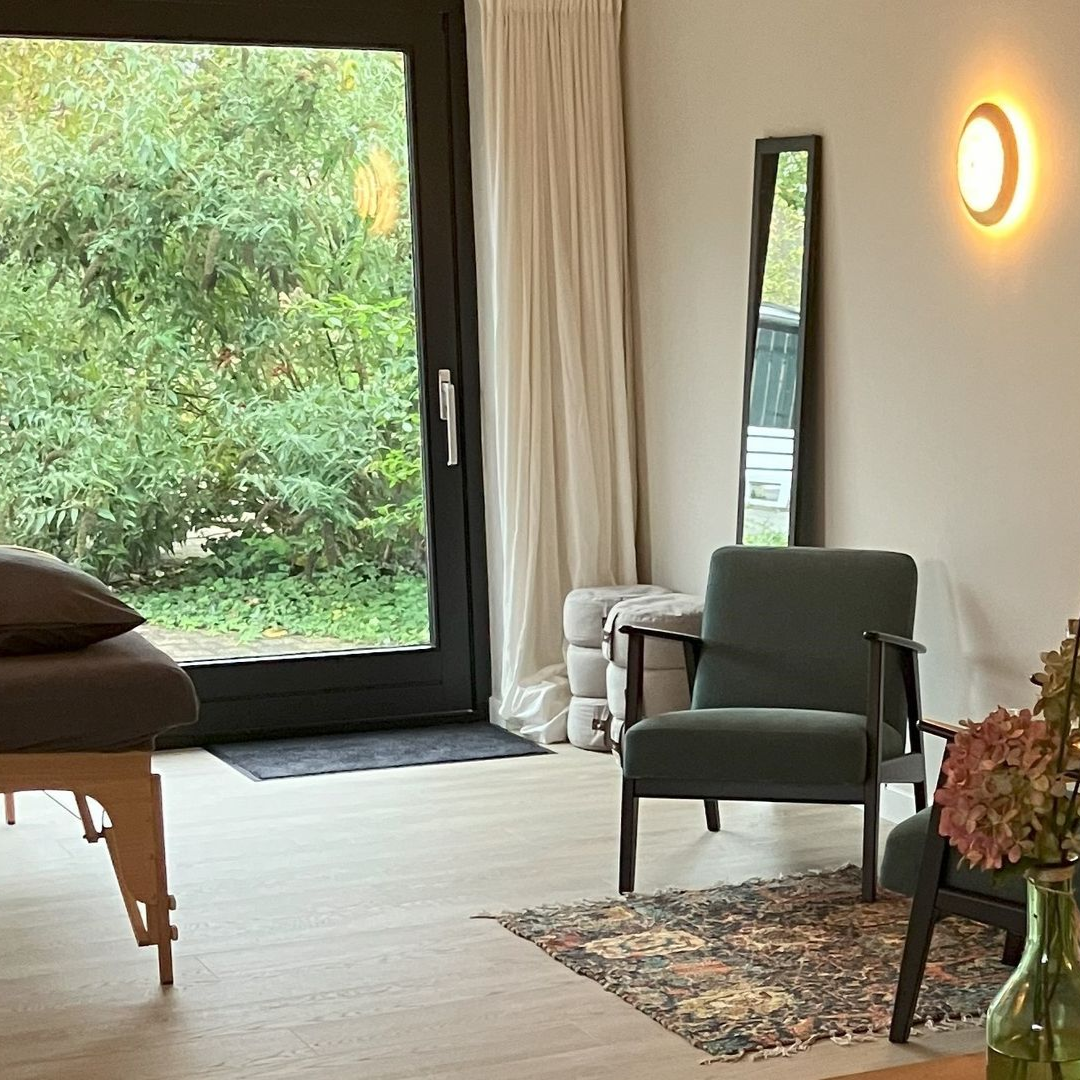 Een kamer met een massagetafel, een fauteuil, een spiegel en een deur met uitzicht op het groen. Warme verlichting.