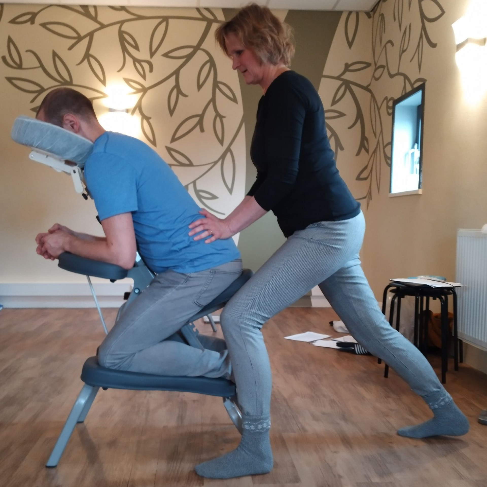 Persoon die een massage zittend ontvangt; therapeut drukt op de rug. Lichtgekleurde kamer, bladerige muur.