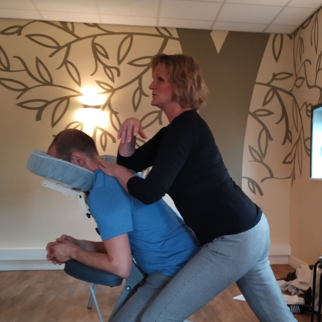 Vrouw geeft een massage aan een persoon die op een massagestoel zit. Lichtgekleurde muren, kunstwerken.