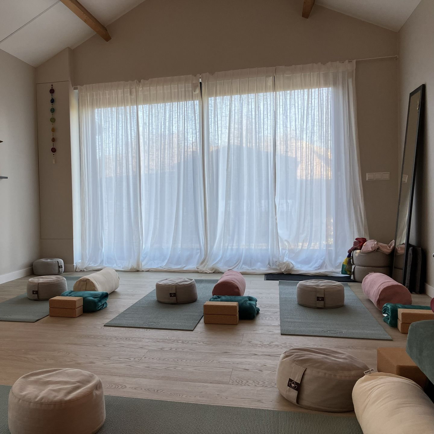 Interieur van een yogastudio met matten, kussens, blokken en vloerkussens, vlak bij een groot raam met transparante gordijnen.