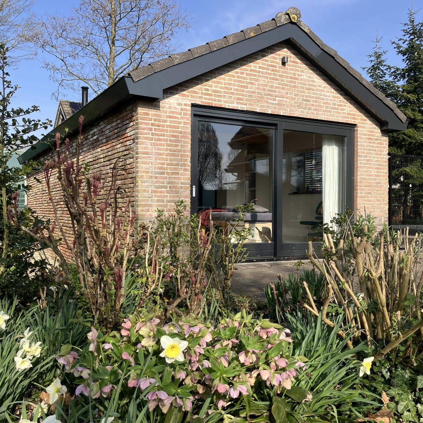 Een stenen huisje met een glazen schuifdeur, omgeven door een tuin met bloemen en struiken.