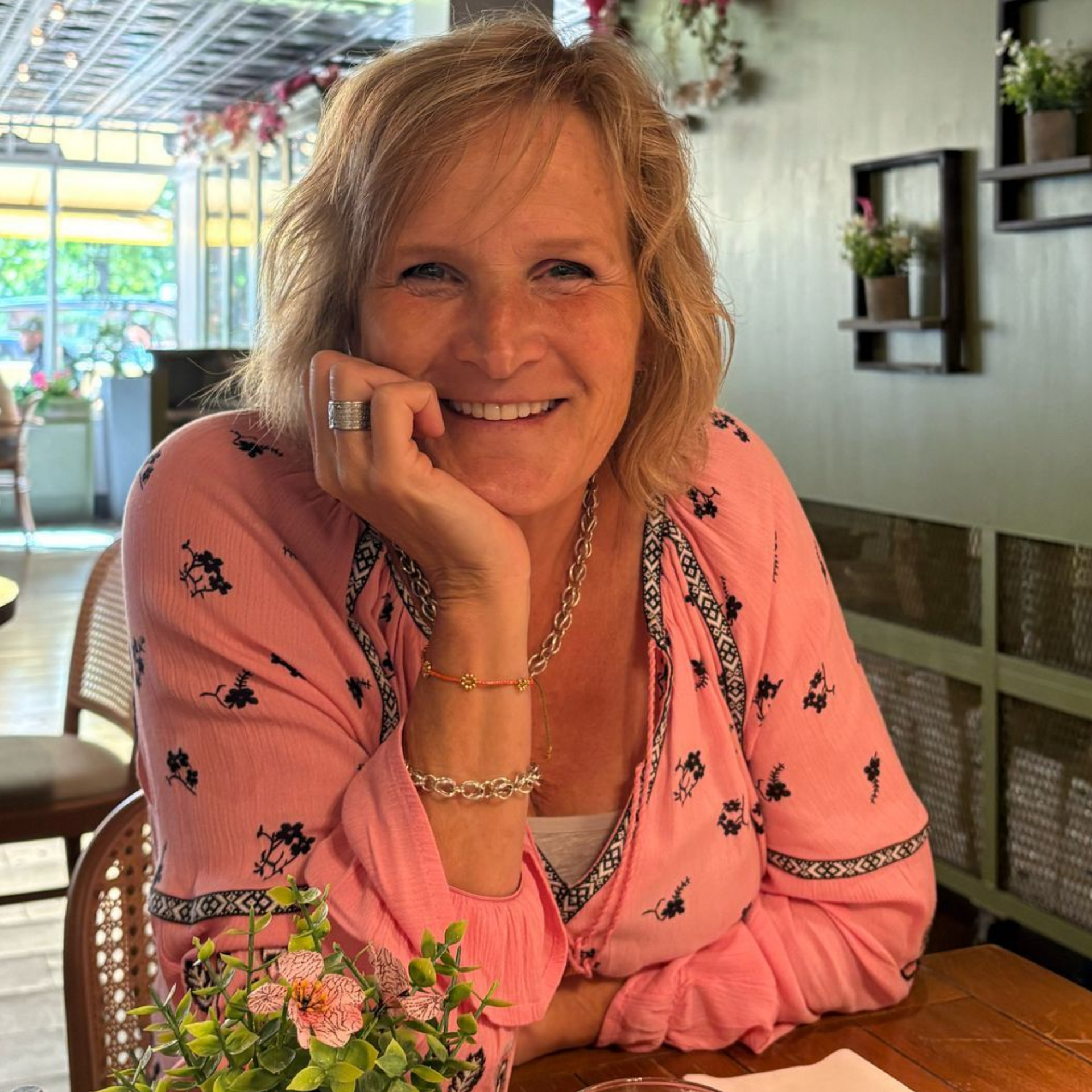 Een vrouw in een roze shirt (eigenaar Senti) lacht en legt haar kin op haar hand aan een tafel met bloemen.