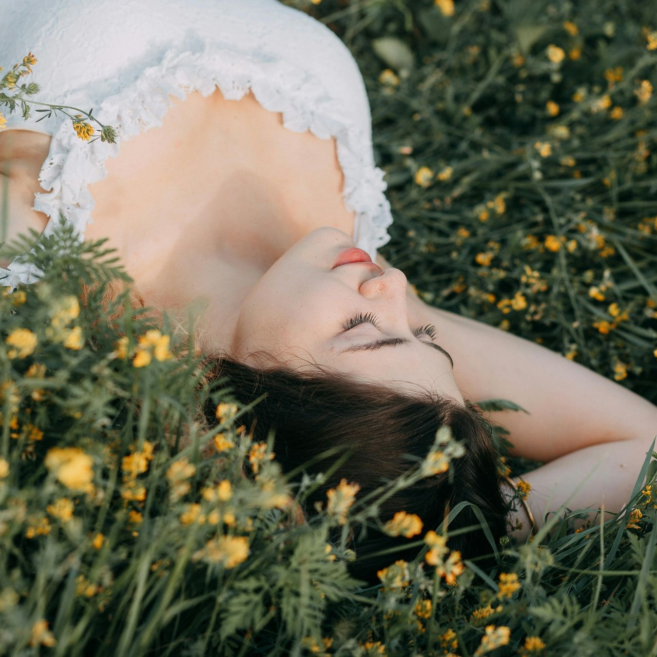 Vrouw in witte jurk ligt in een veld met gele bloemen, ogen gesloten, ontspannen uitdrukking.