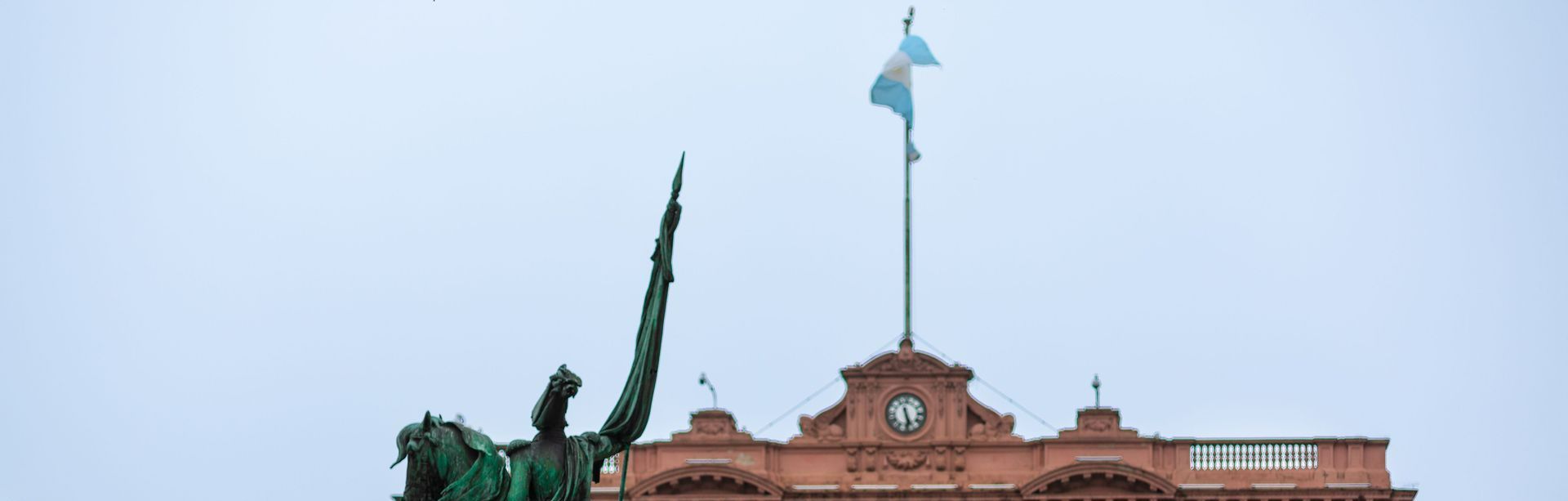 Uma estátua de um homem montado num cavalo em frente a um prédio com uma bandeira tremulando ao vento.