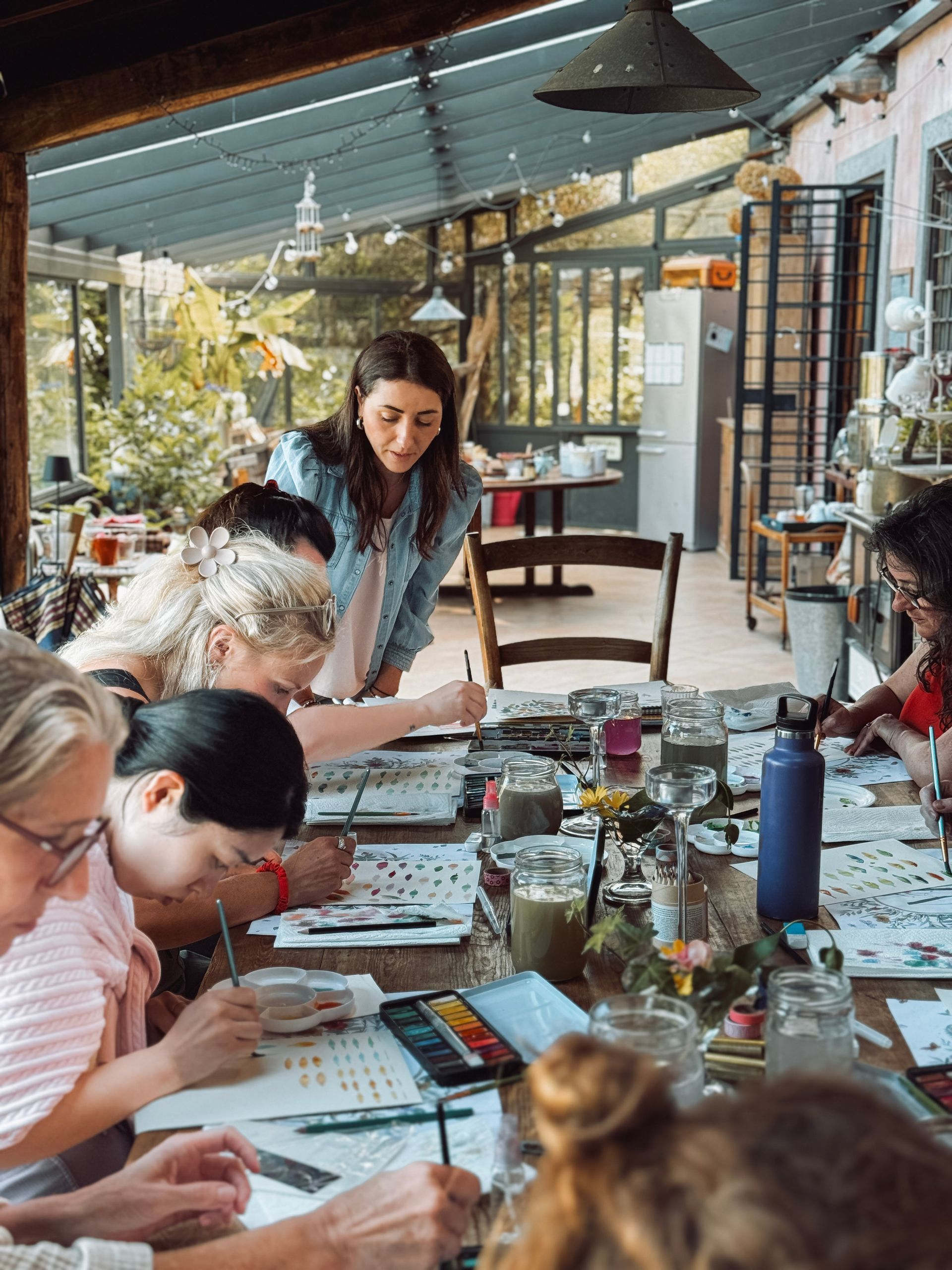 People painting at a table indoors. A woman looks over their shoulders, offering instruction. The setting is bright and airy.