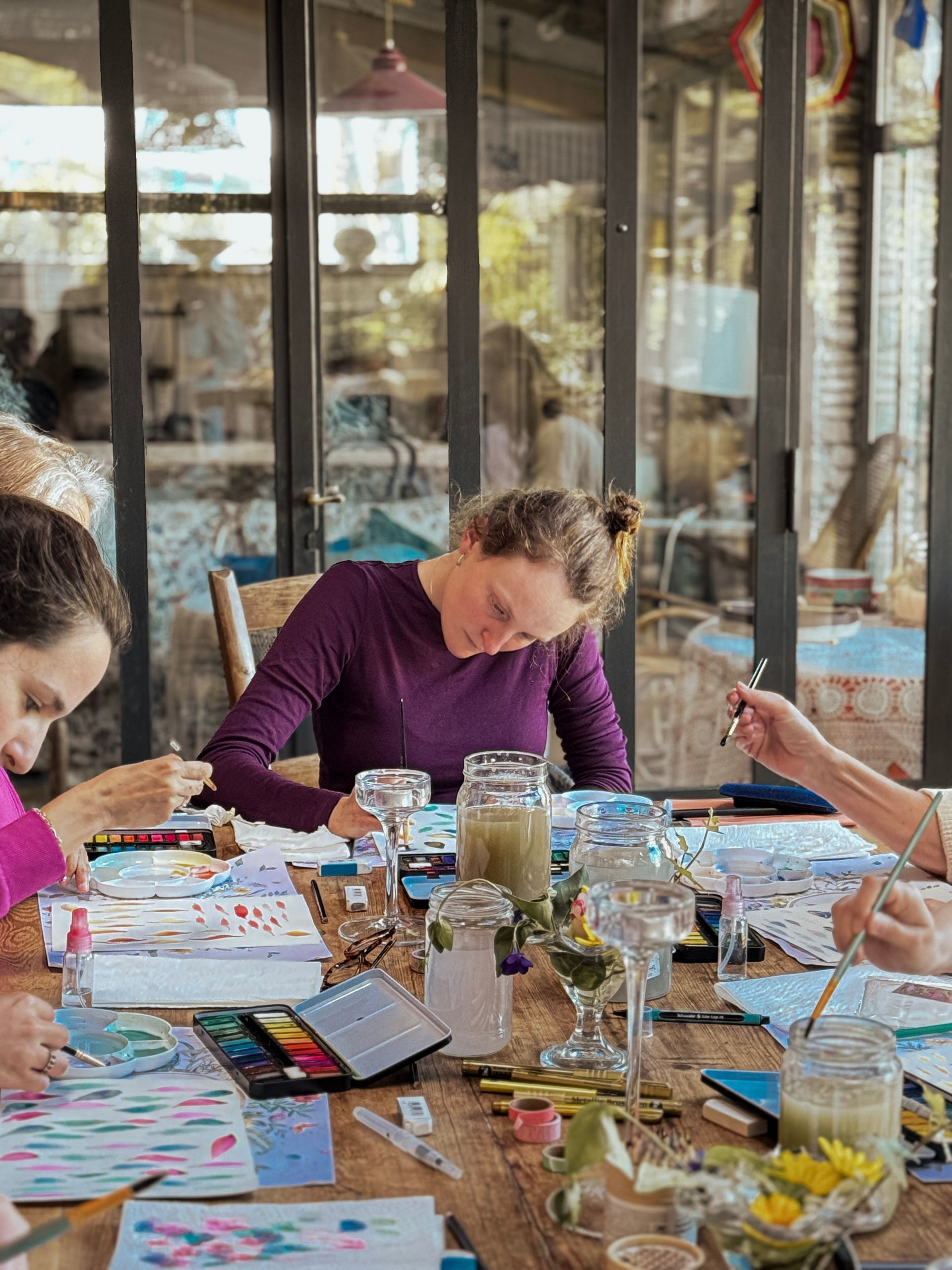 People painting at a table indoors. A woman in purple concentrates on her art. Paint supplies and glasses are visible.