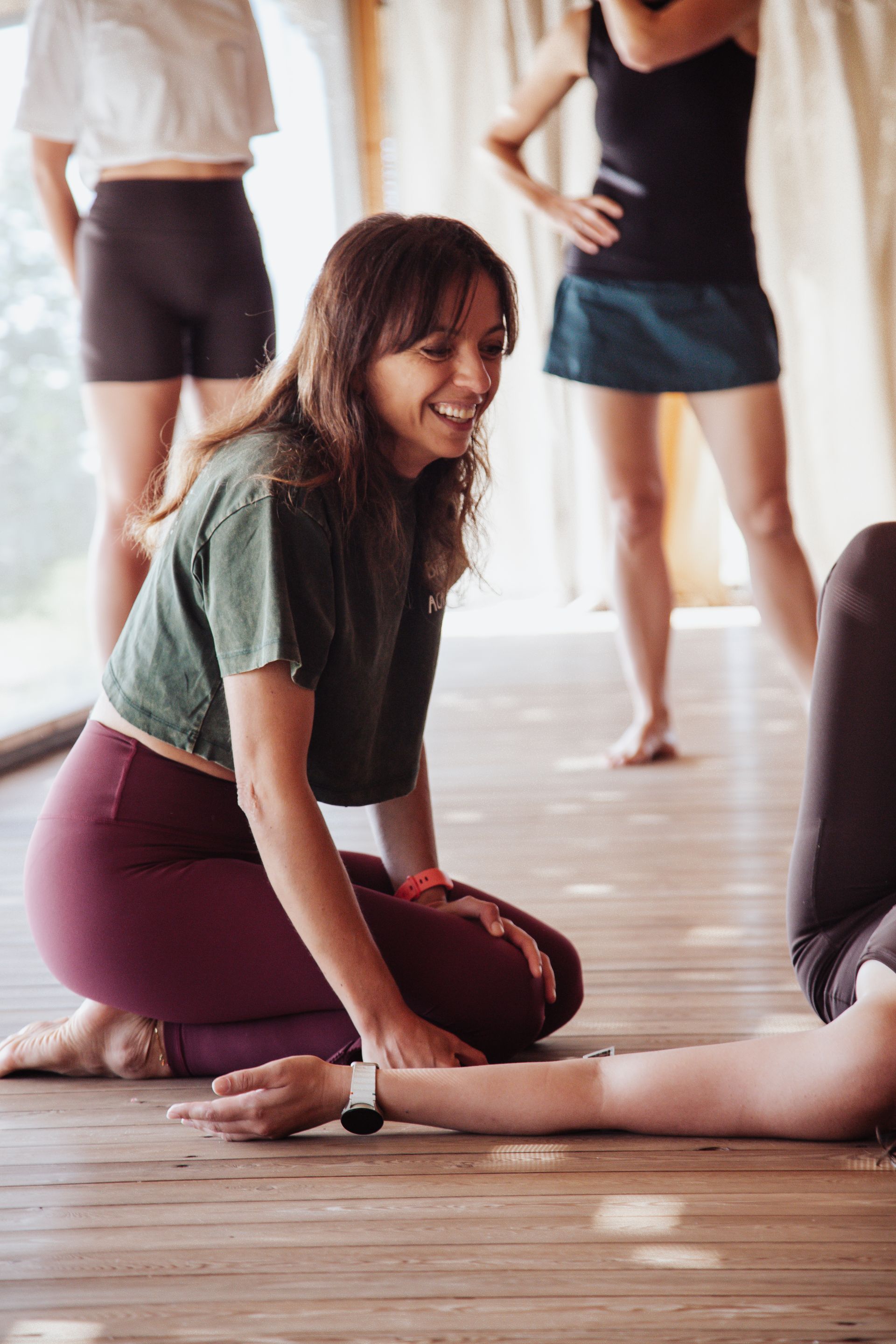 Woman smiling, assisting another with a yoga pose on a wooden floor, other people in the background.