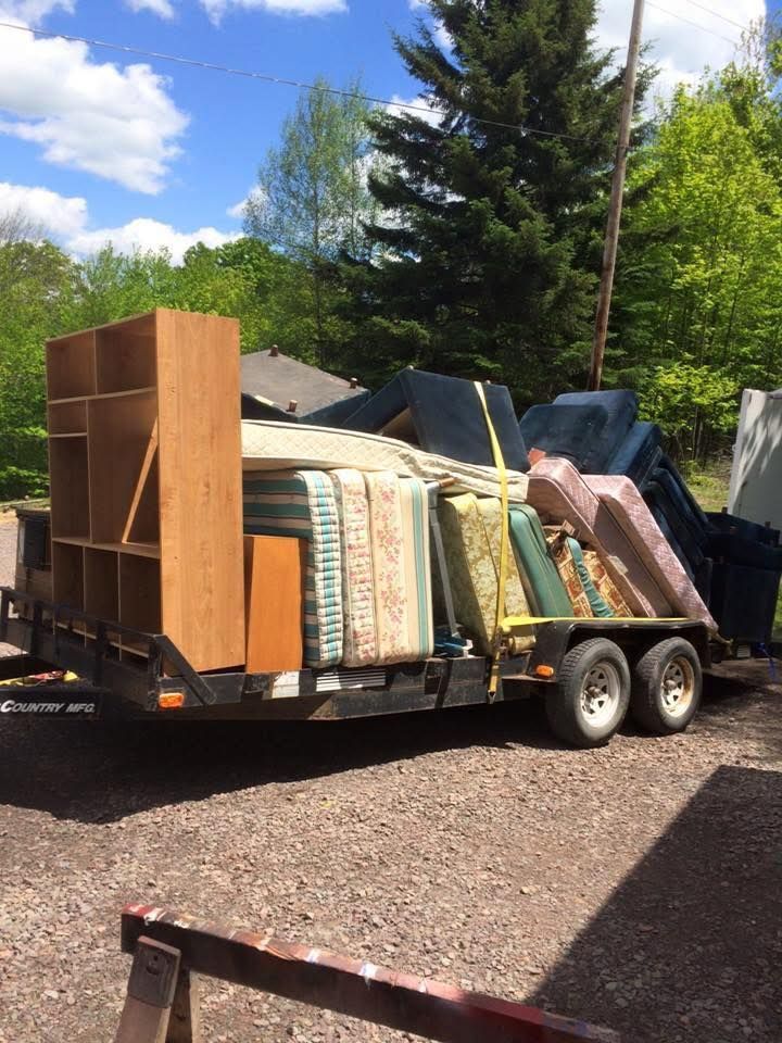A trailer filled with furniture and mattresses is parked in a gravel lot.