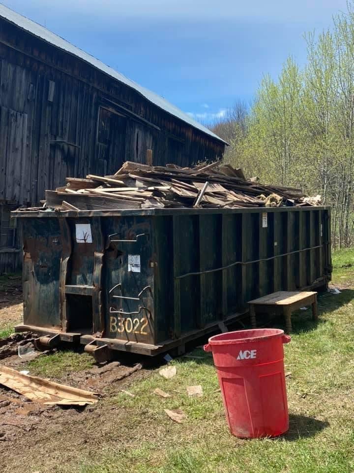 A large dumpster filled with wood is sitting in front of a barn.