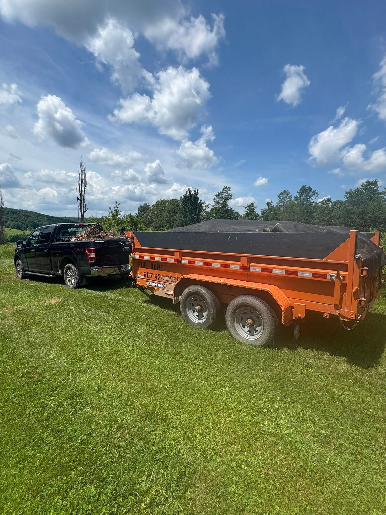 A black truck is towing an orange dump truck in a grassy field.