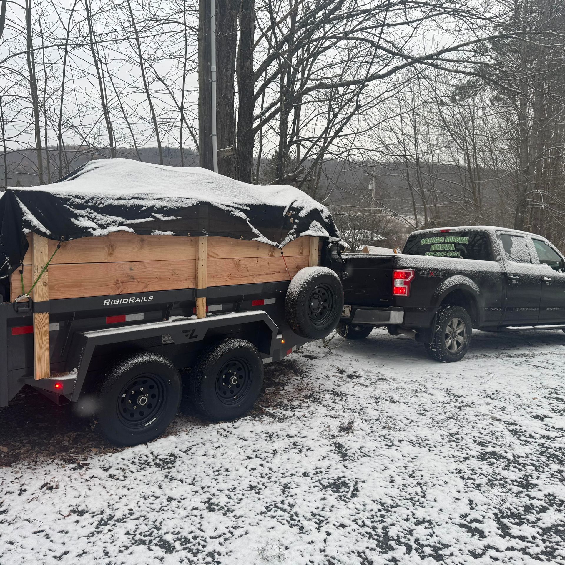 Black pickup truck towing a trailer with wooden sides, covered in snow, in a snowy outdoor setting.