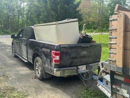 Black pickup truck hauling a large white appliance on a trailer in a wooded roadside area
