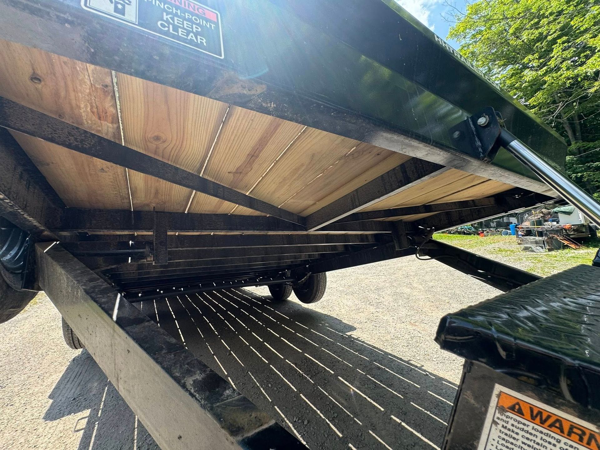 Underside of a black trailer with wooden floor panels, viewed from below in a gravel lot.