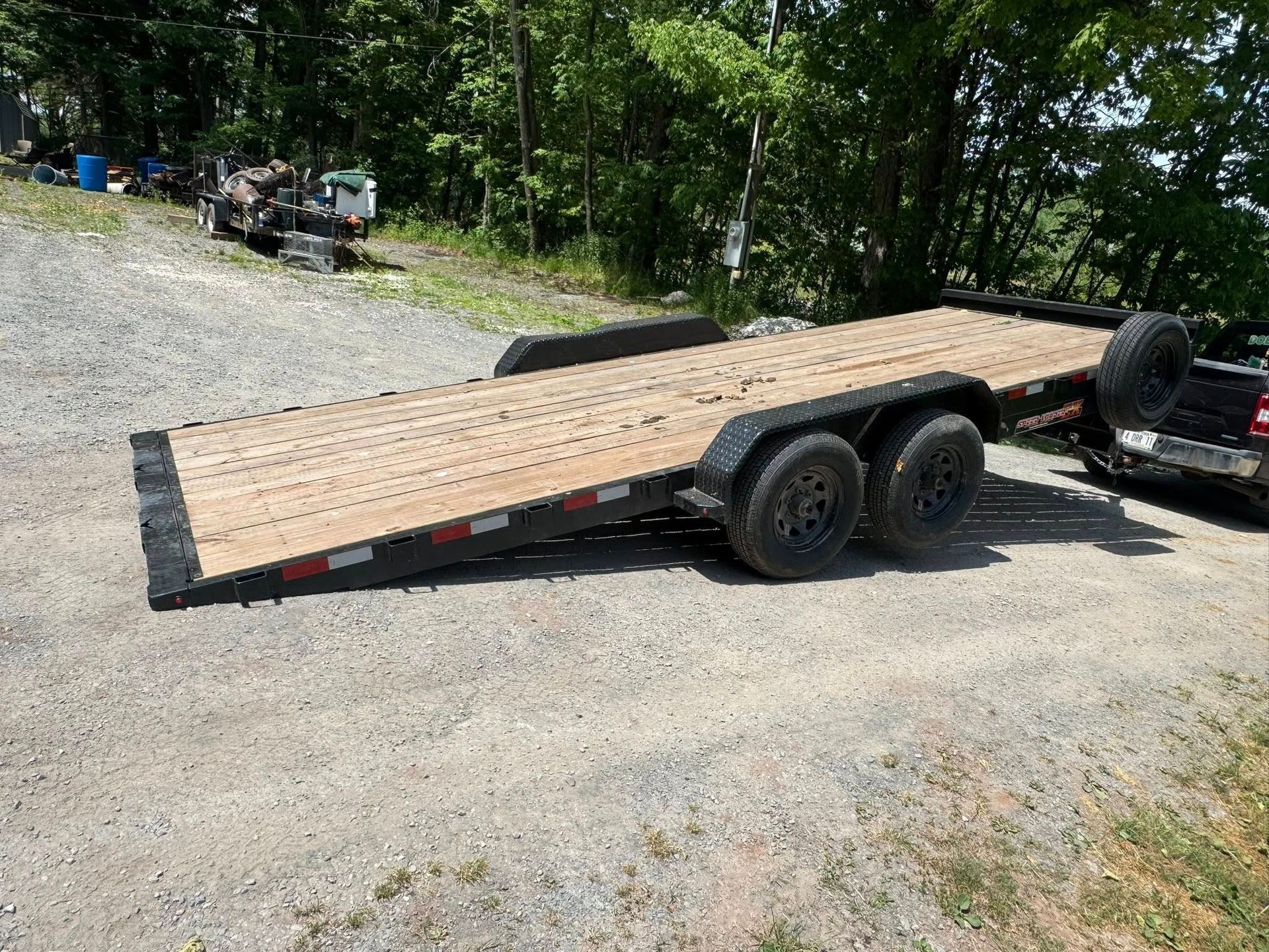 Black flatbed car trailer parked on a gravel lot near trees, with wooden deck and dual axles.