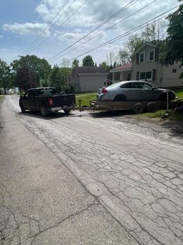 Black truck towing a car on a trailer down a cracked road in front of houses.