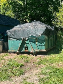Green dumpster covered with black netting in a grassy area, near a wooden building.