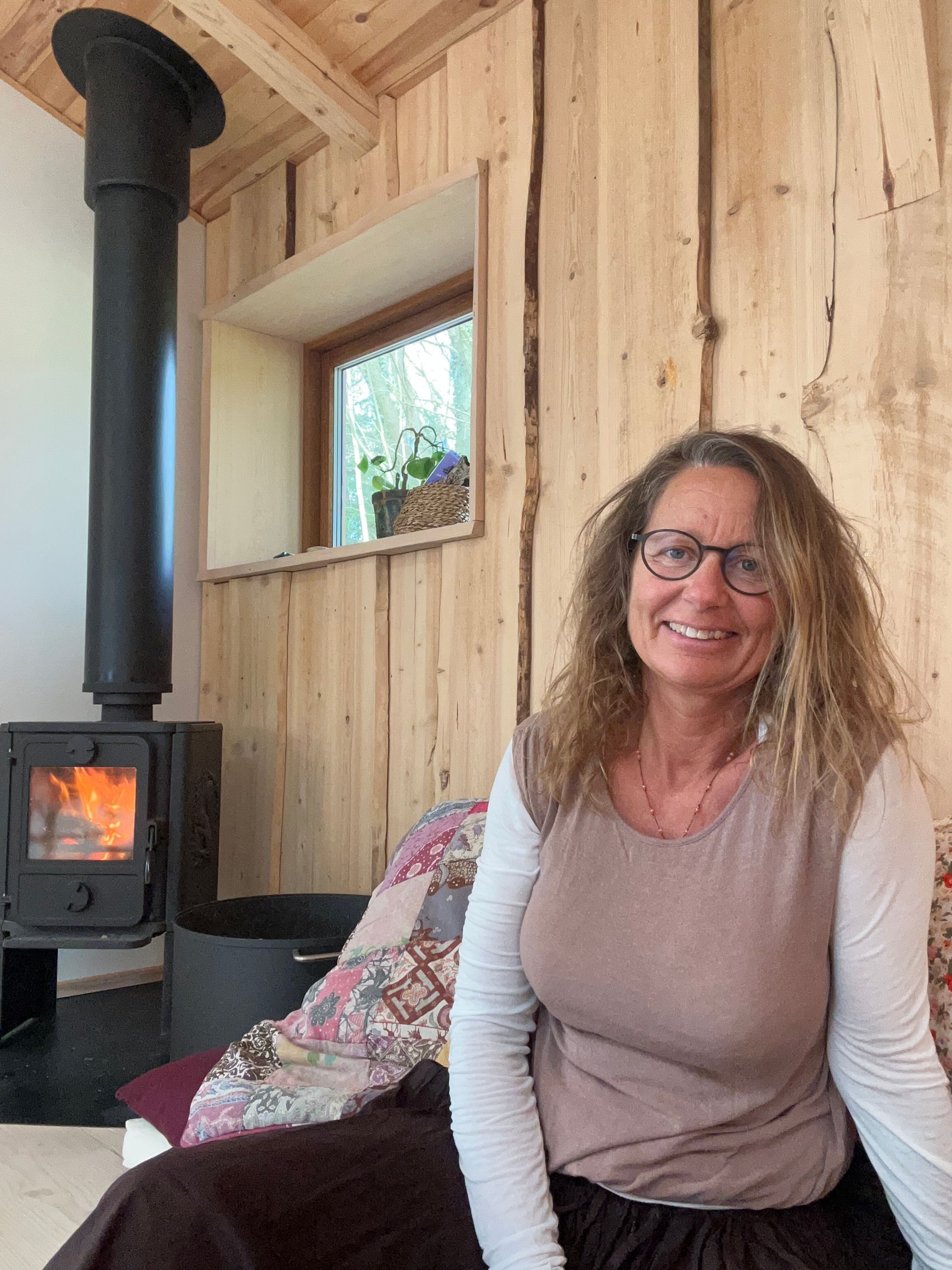 En smilende person med briller sidder foran en tændt brændeovn i et rum med lyse træpaneler på væggene.