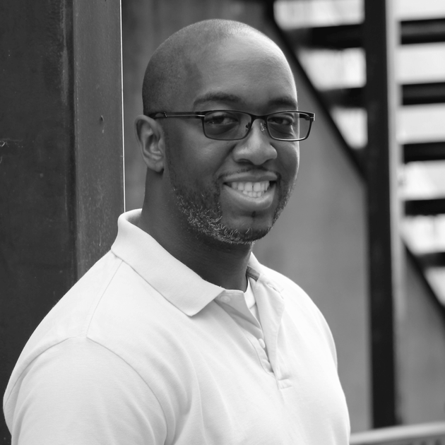 A man with a short beard and glasses smiling while leaning against a dark wall.