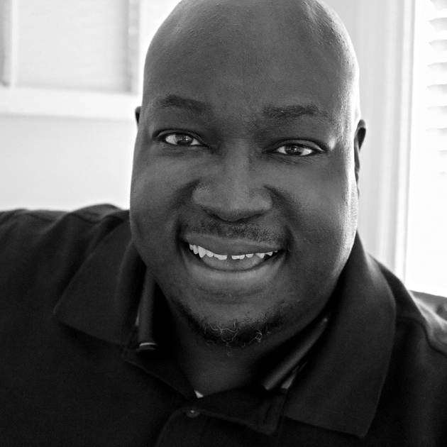 Close-up, black-and-white portrait of a smiling person with a shaved head wearing a polo shirt.