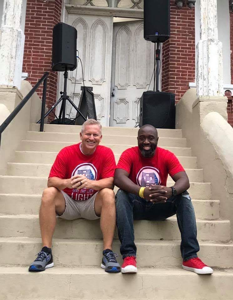 Two people in red shirts sitting on concrete steps in front of a brick building with two large speakers.