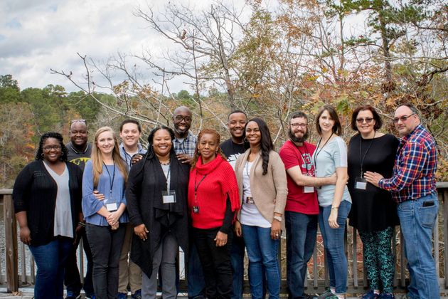 A group of fourteen people stand together smiling outdoors in front of trees, posing for a photo on a deck.
