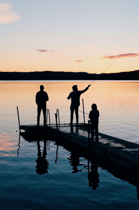 Silhouetted figures fishing from a dock at sunset over a calm lake.