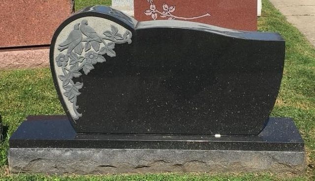 A black gravestone with a bird and flowers on it is in a cemetery.